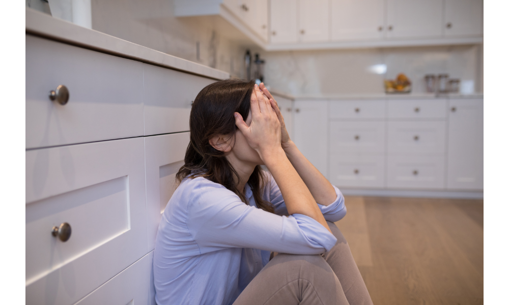 Mulher triste sentada no chão de uma cozinha.