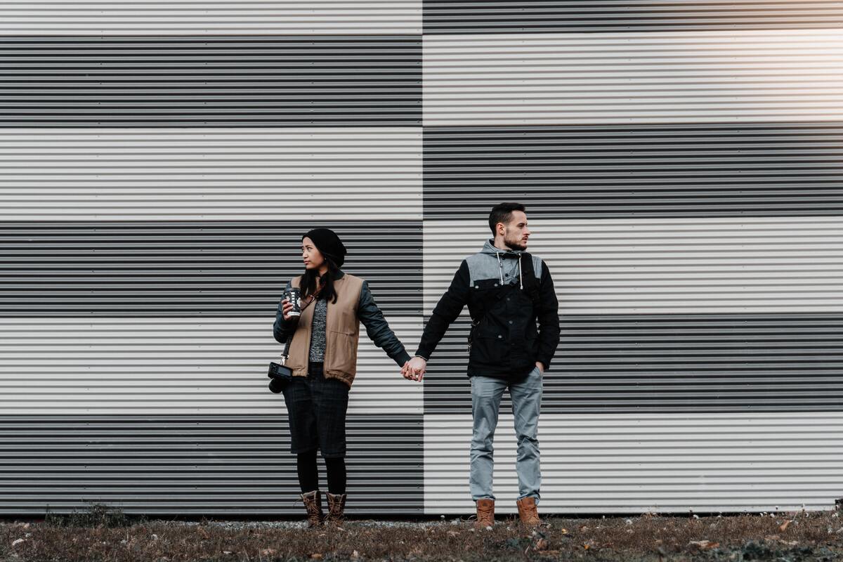 Casal em pé, de mãos dadas, olhando para lados opostos.