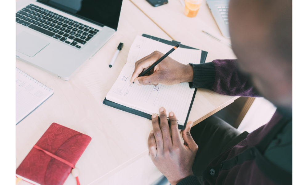 Homem se organizando com um caderno e um notebook ao lado.