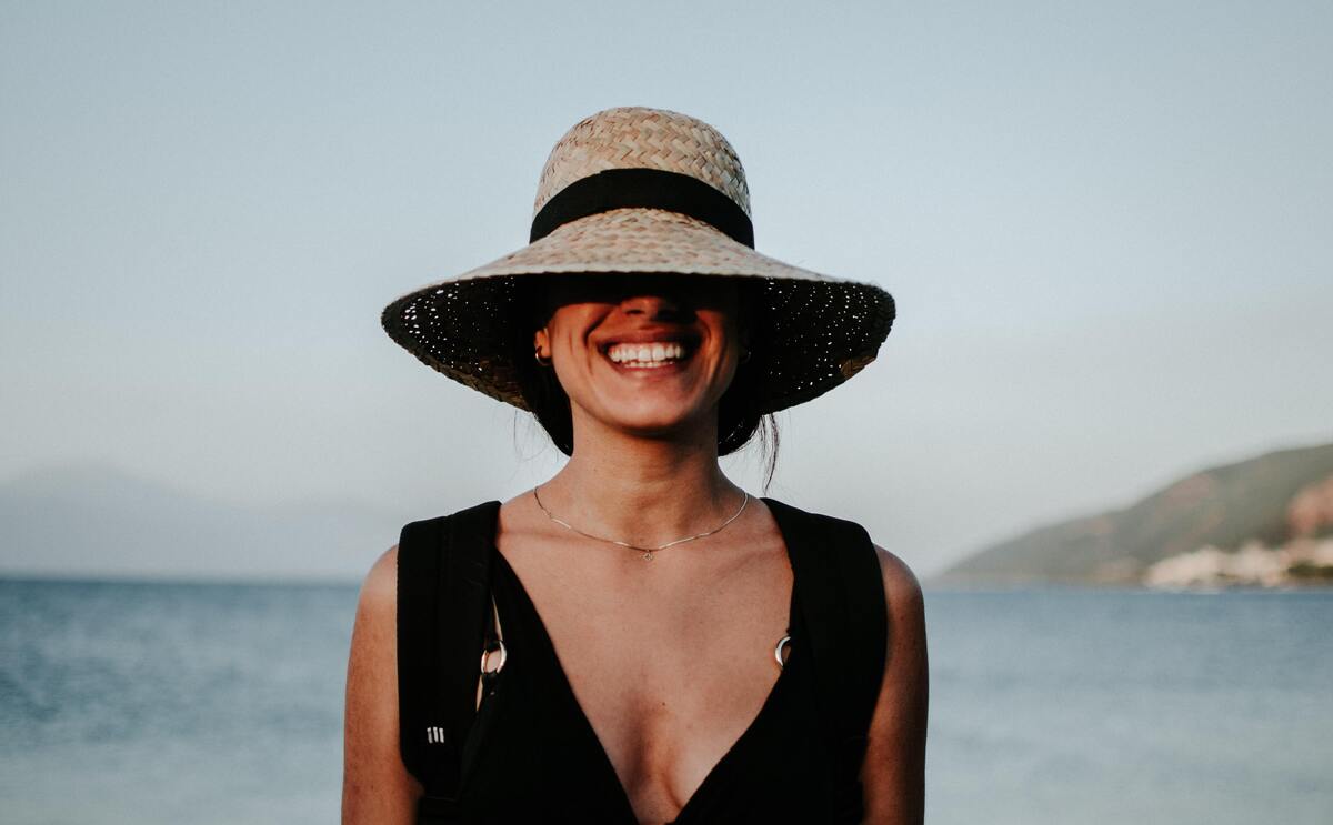 Mulher usando vestido preto, com um chapéu cobrindo os olhos e sorrindo.