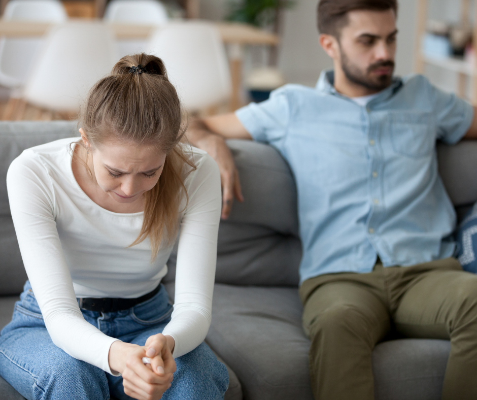 Casal sentado em um sofá, a mulher está com as mãos sob as pernas, enquanto o homem se mostra indiferente