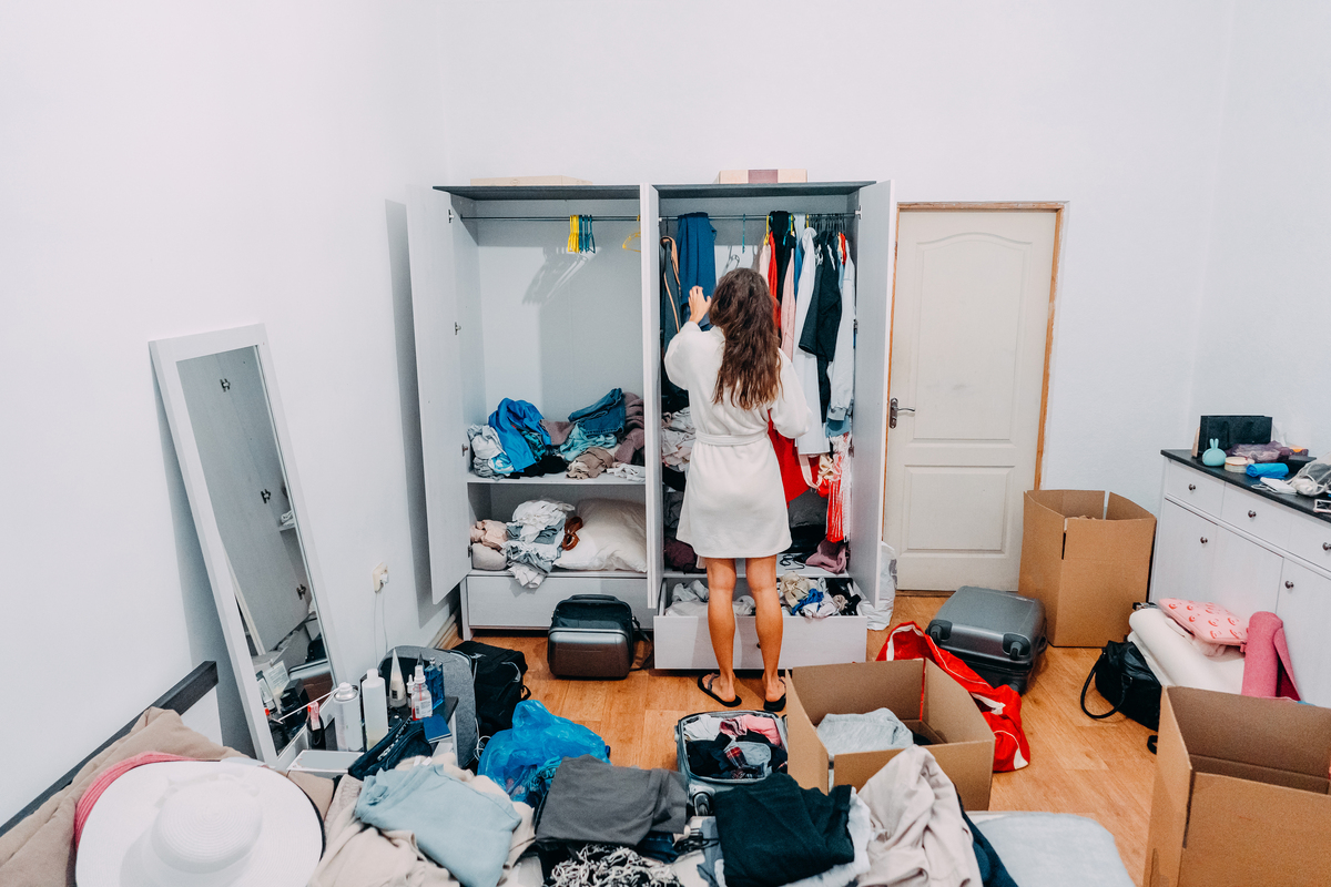 Mulher mexendo em guarda-roupa de casa bagunçada, com diversas roupas e objetos jogados pelo chão e pela cama. 