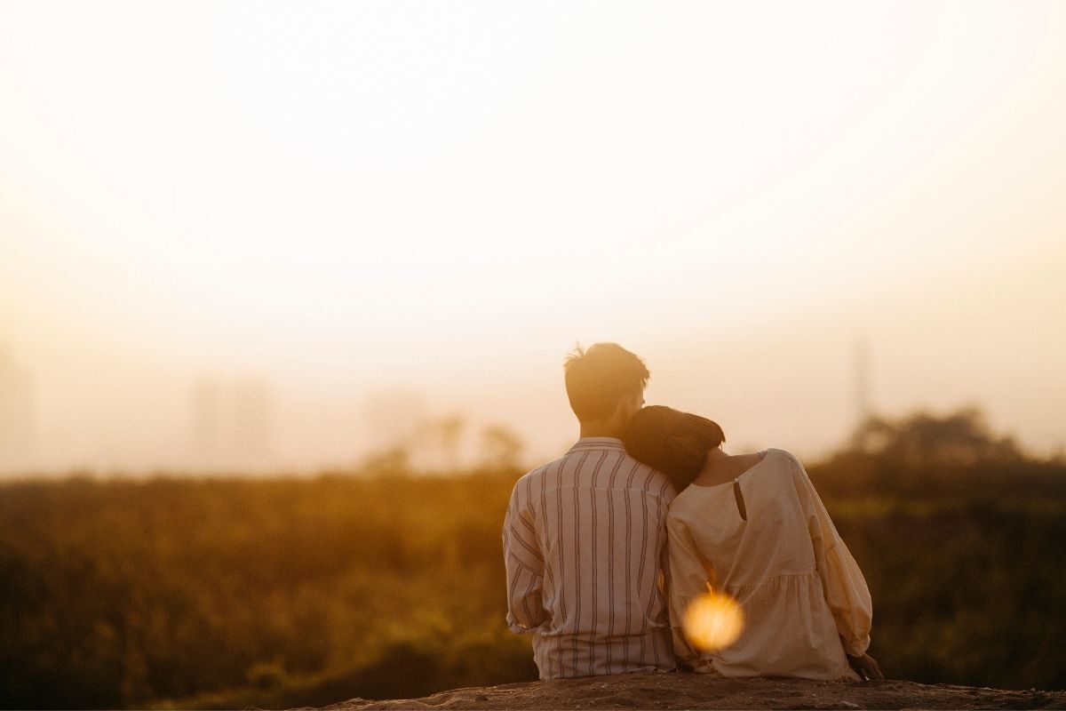 Casal de costas para a foto, abraçados vendo o pôr do Sol em um banco de praça