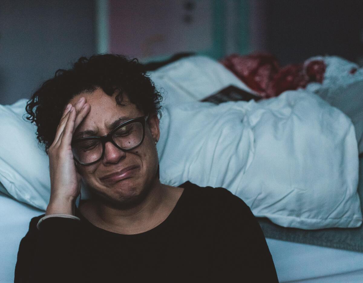 Mulher chorando enquanto alguém dorme na cama ao fundo.