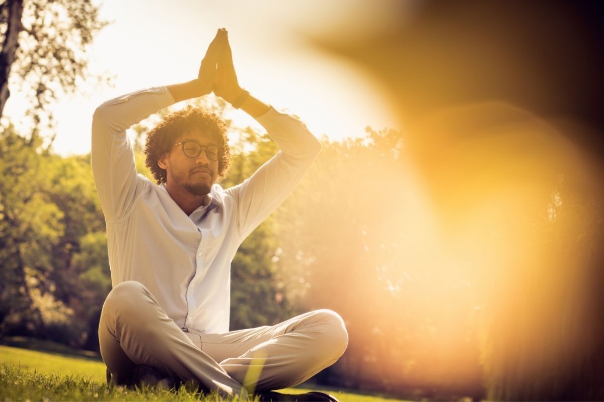 Homem vestido de branco meditando em um parque, com as mãos unidasd em cima da cabeça