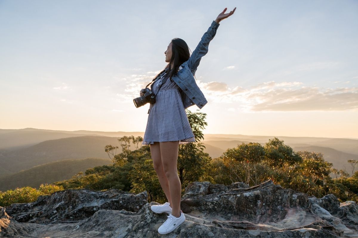 Mulher admirando a paisagem da natureza, se divertindo com um dos braços para o alto e uma camêra fotográfica