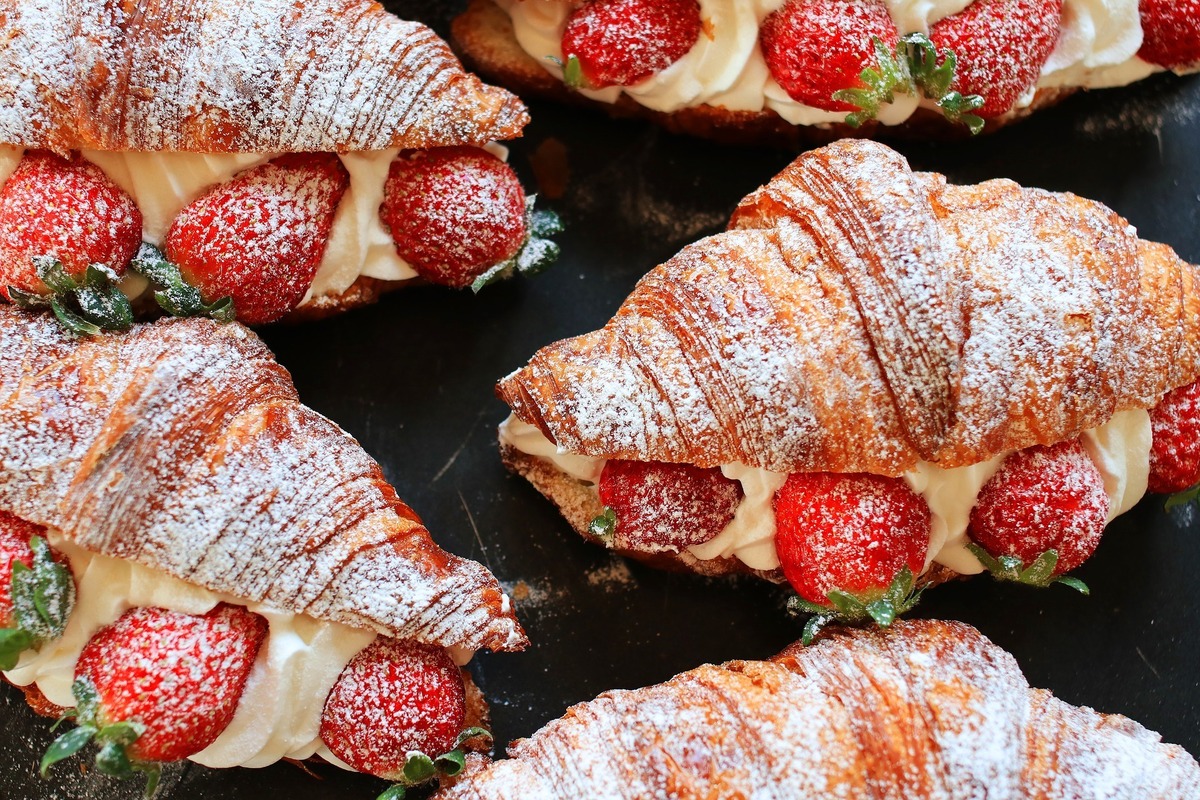 Vários croissants, um tipo de pão doce, bem recheados com creme branco e morangos, além de estarem cobertos por açúcar de confeiteiro.