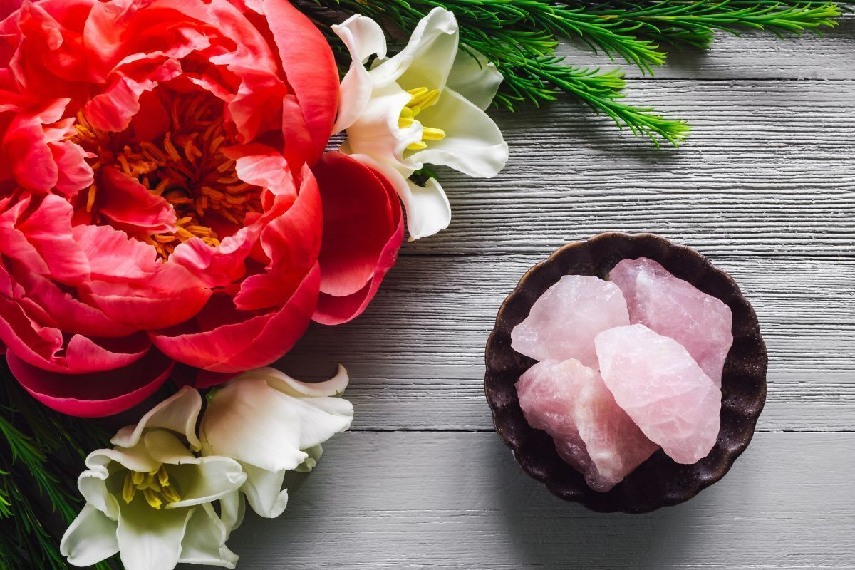 Vasilha com cristais pequenos de quartzo rosa em uma mesa de madeira. Na decoração, peônia vermelha e tulipas amarelas.