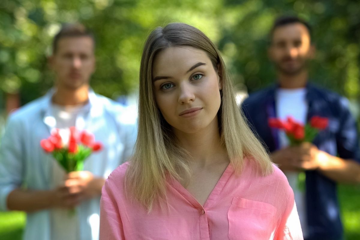 Mulher com olhar de dúvida. Atrás dela há dois homens com buquê de rosas nas mãos