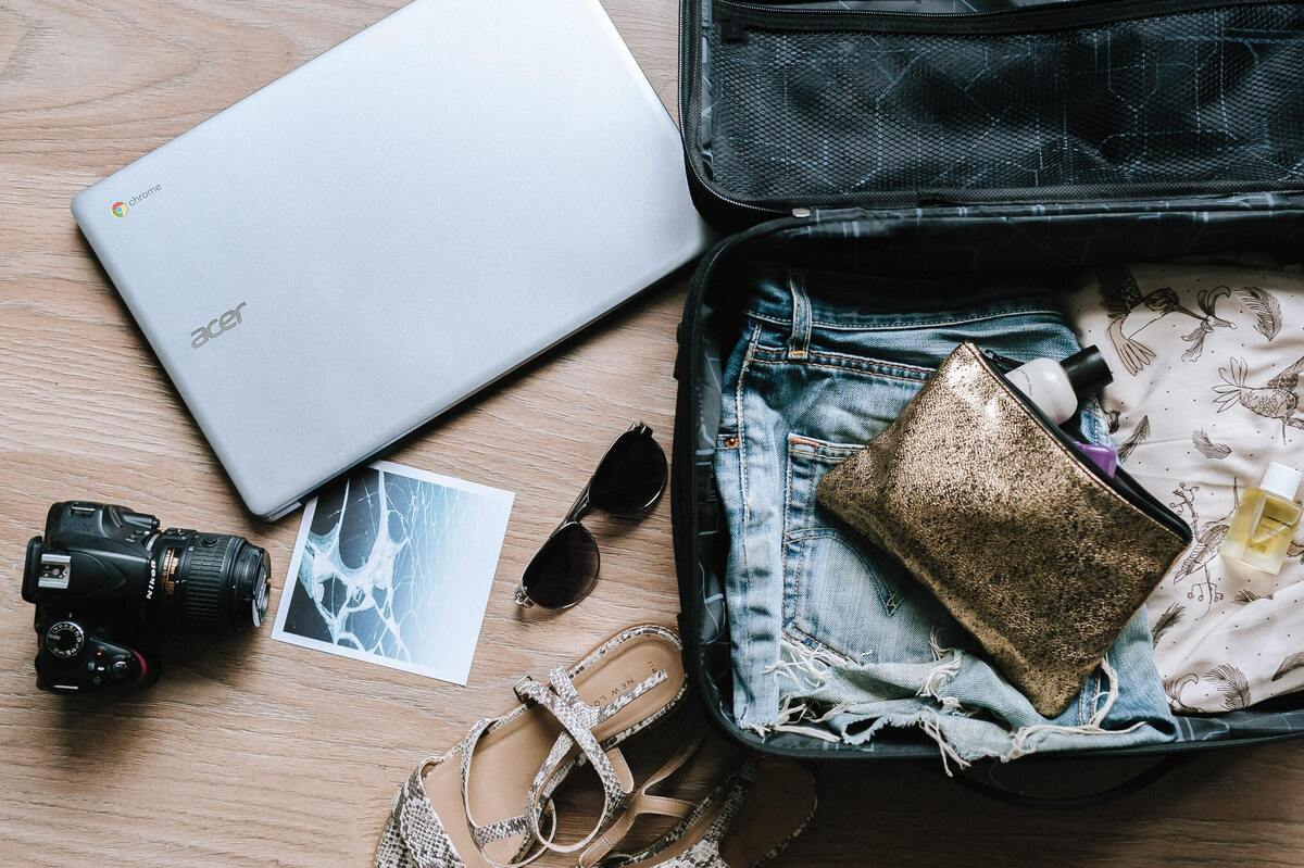 Mala aberta com algumas roupas dentro e, do lado de fora, um notebook, uma câmera, óculos de sol, um par de sapatos e uma fotografia.