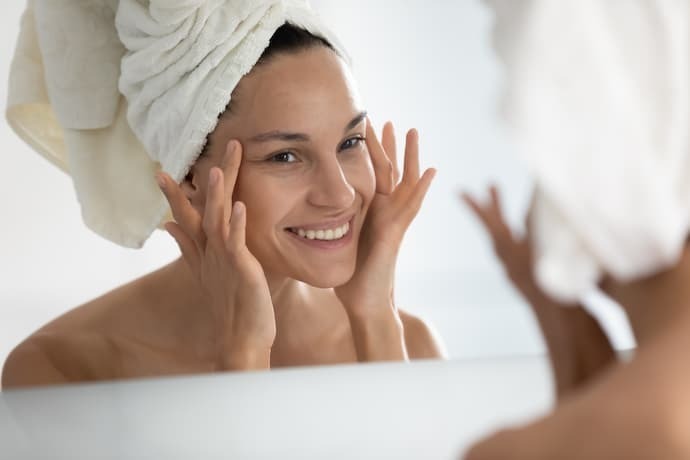 Mulher sorridente se vendo no espelho enquanto passa creme no rosto