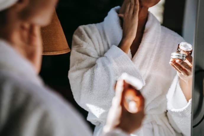 Reflexo de mulher se olhando no espelho e segurando pote com creme