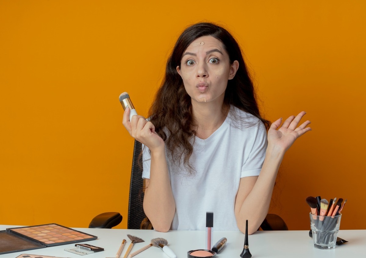 Mulher atrás de uma mesa com maquiagens com expressão de dúvida segurando um pote de base para o rosto