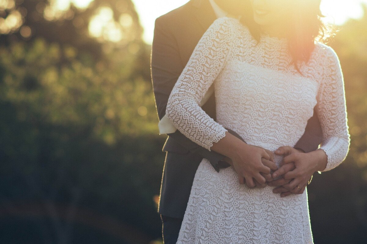 Homem de terno abraçando mulher de vestido branco por trás, representando o relacionamento de Câncer e Gêmeos dependendo do gênero de cada um.