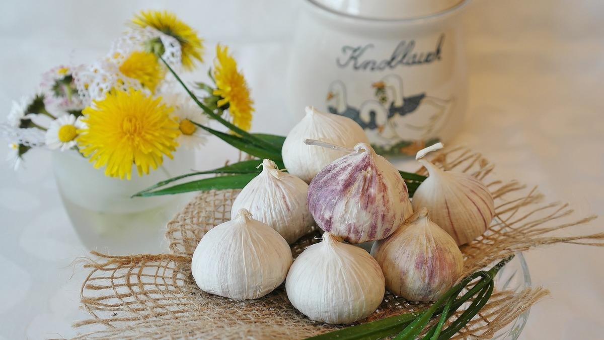 Diversas cabeças de alho organizadas em cima de pequeno cesto de palha, ao lado de vaso com flores amarelas.