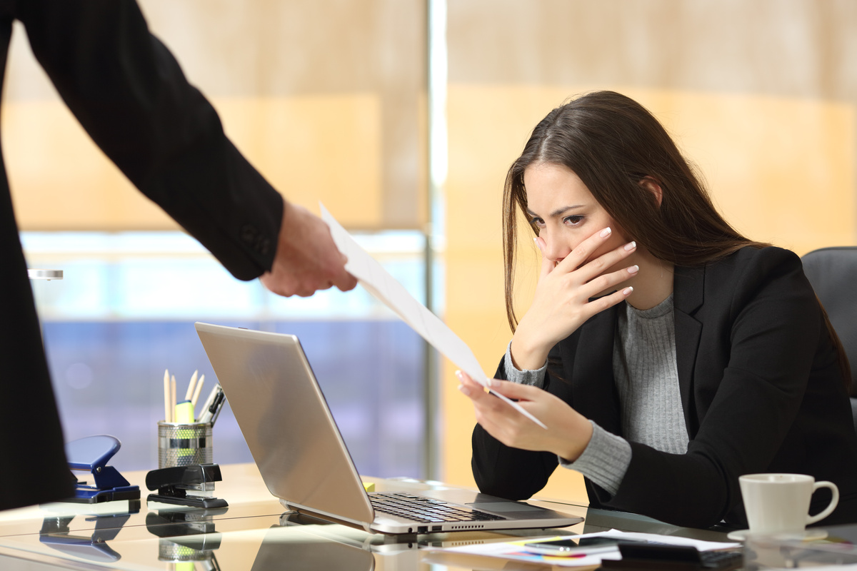 Mulher sentada na mesa, com expressão surpresa ao receber papéis anunciando sua demissão.