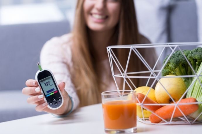 Mulher segurando medidor de glicemia com copo de suco e cesta de frutas na sua frente