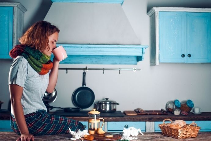 Mulher resfriada sentada na mesa de uma cozinha tomando um chá