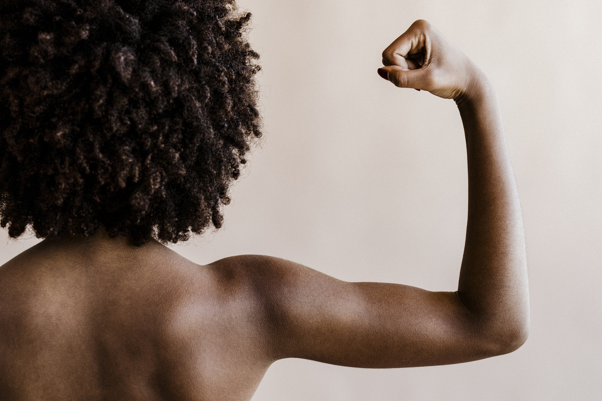 Mulher negra, de cabelos castanhos e cacheados, de costas estendendo o braço direito.