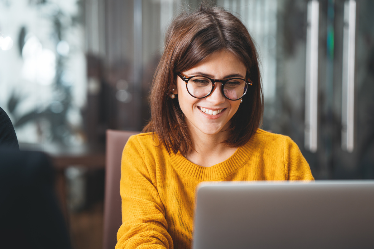 Mulher branca de óculos e suéter amarelo trabalhando com notebook aberto.