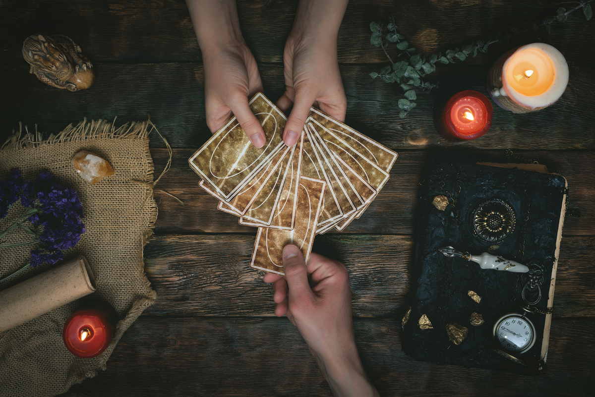 Duas pessoas sentadas de frente para a outra em mesa, envoltas de velas e outros objetos. Uma segura um monte de cartas de Tarot viradas para baixo, enquanto a outra retira uma carta desse monte.