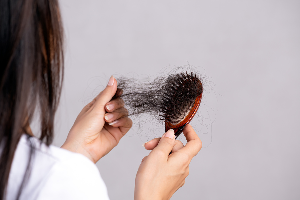 Mulher de costas segurando escova de cabelo e arrancando dela tufos de cabelo preto caído.