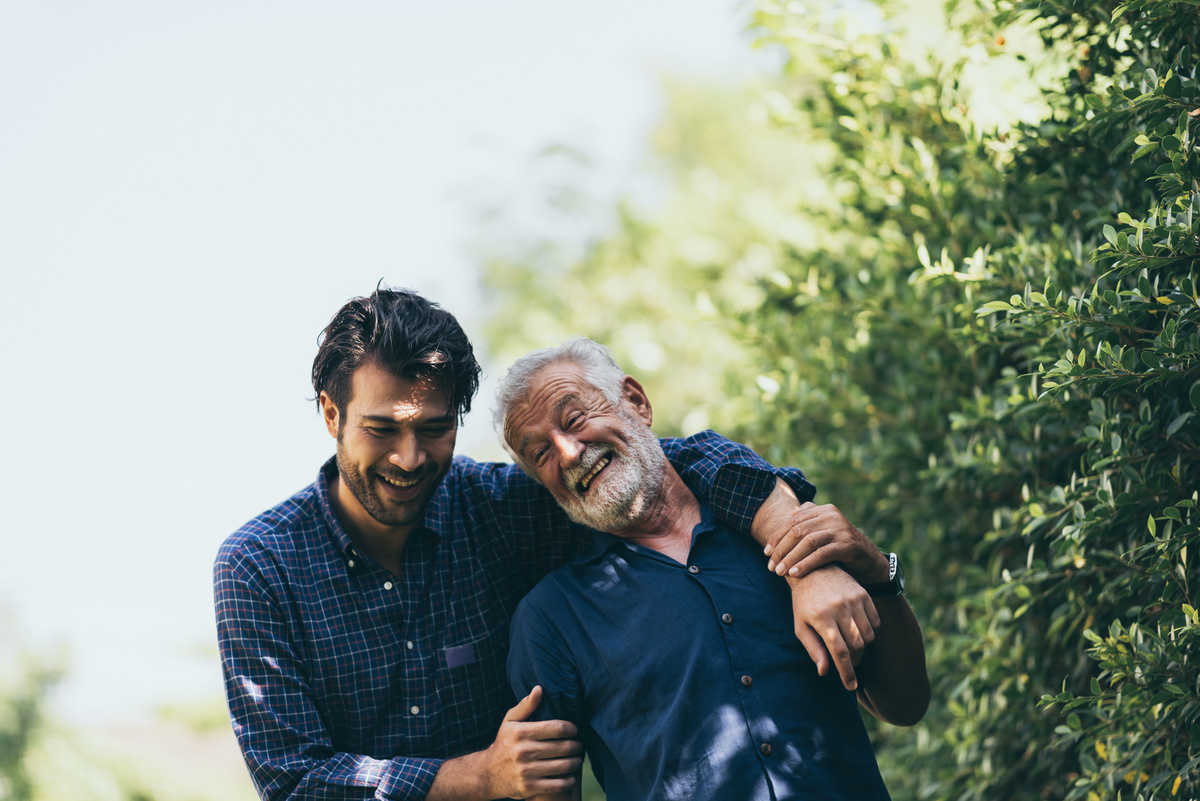 Homem caminhando pelo campo enquanto abraça os ombros de senhor e ambos sorriem, simbolizando sonhar com pai falecido.