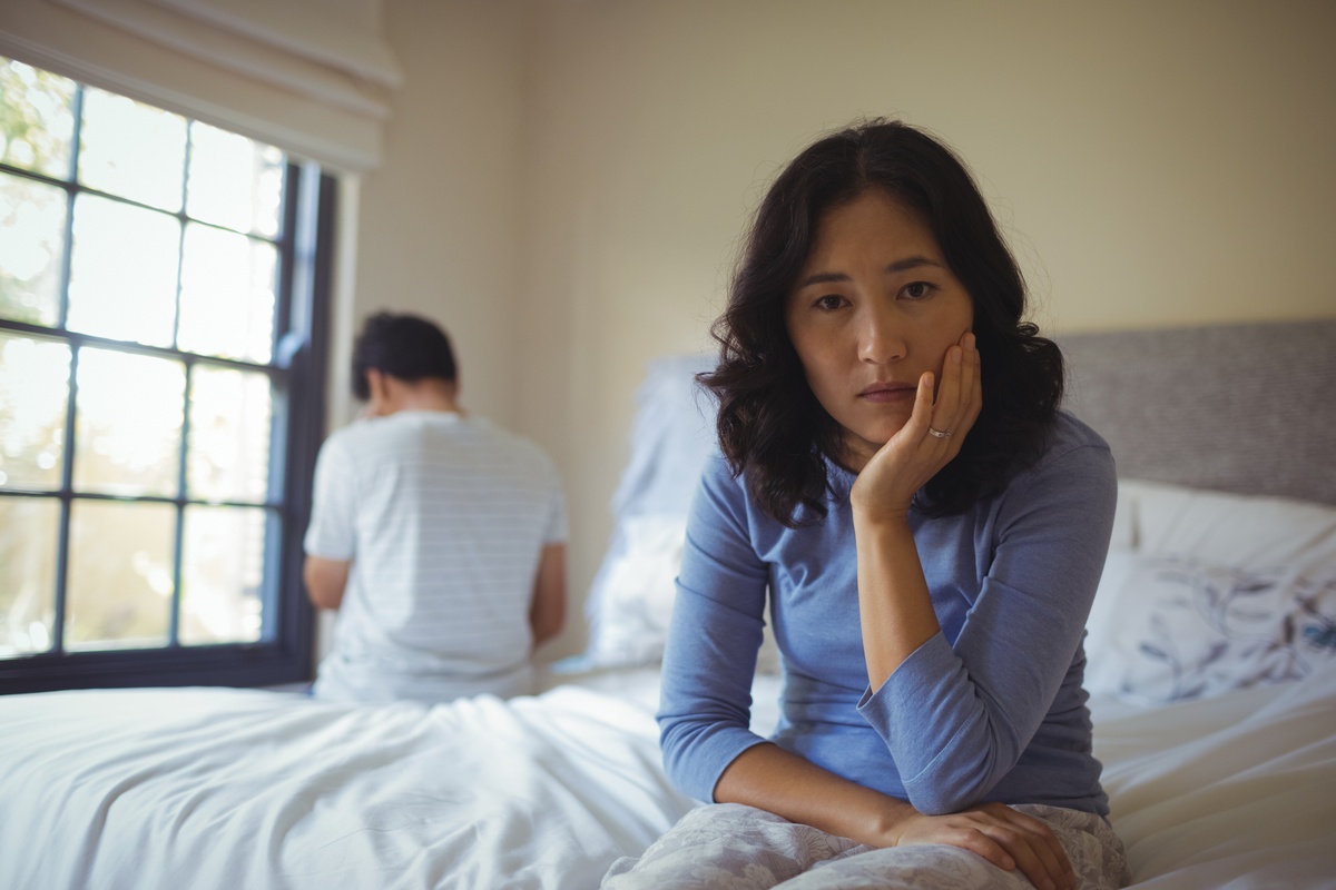 Casal de homem e mulher sentados nos dois extremos da cama, com o homem de costas no segundo plano, e mulher à frente, portando uma expressão triste.