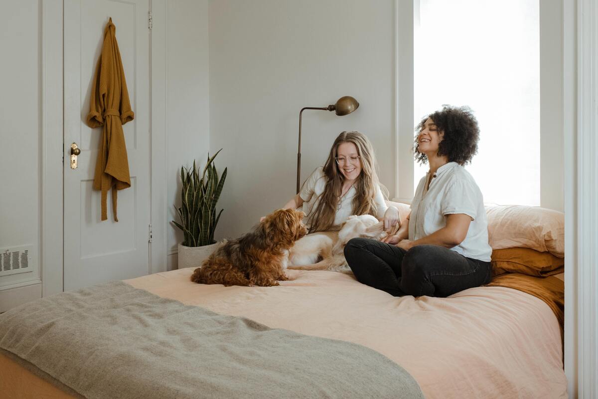 Duas mulheres sentadas numa cama com cachorro ao lado.