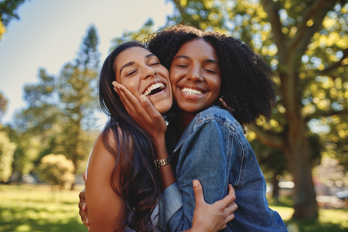 Duas amigas em meio à natureza, se abraçando de olhos fechados e sorrindo, representando a amizade entre Gêmeos e Peixes.