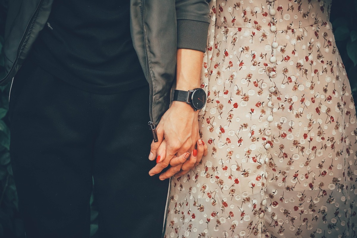 Mulher usando um vestido florido, enquanto tem as unhas pintadas de vermelho, dando as mãos para homem, que usa um relógio de pulso e roupas pretas, representando Peixes e Gêmeos.