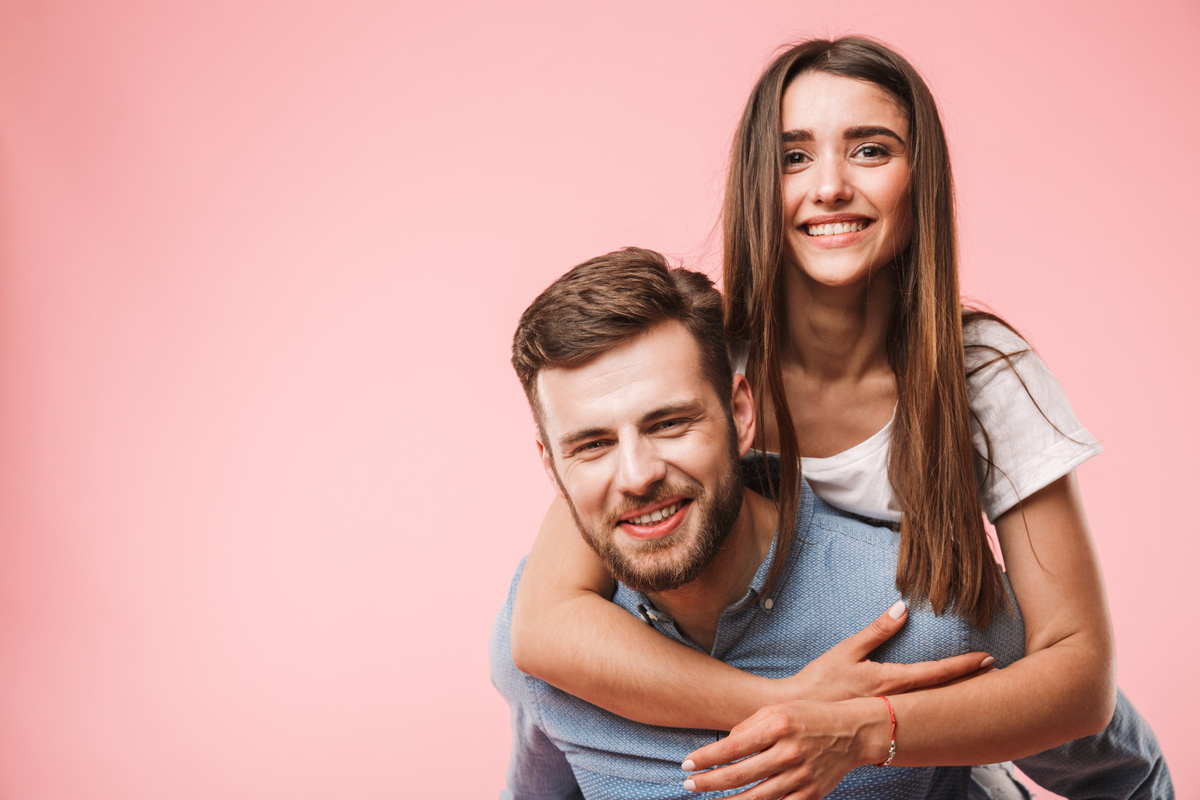 Em fundo rosa, homem carregando mulher nas costas enquanto ambos sorriem, representando Virgem e Escorpião de acordo com o gênero.