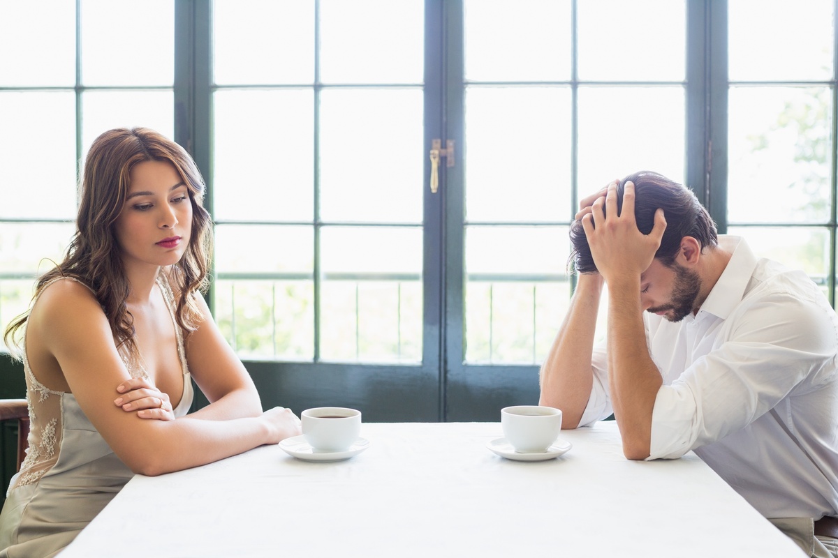 Homem e mulher sentados em mesa, um de frente para o outro, com xícaras de café em frente a eles. A mulher tem o olhar distante, enquanto o homem tem as mãos na cabeça e o rosto abaixado, representando sinais de amarração amorosa.