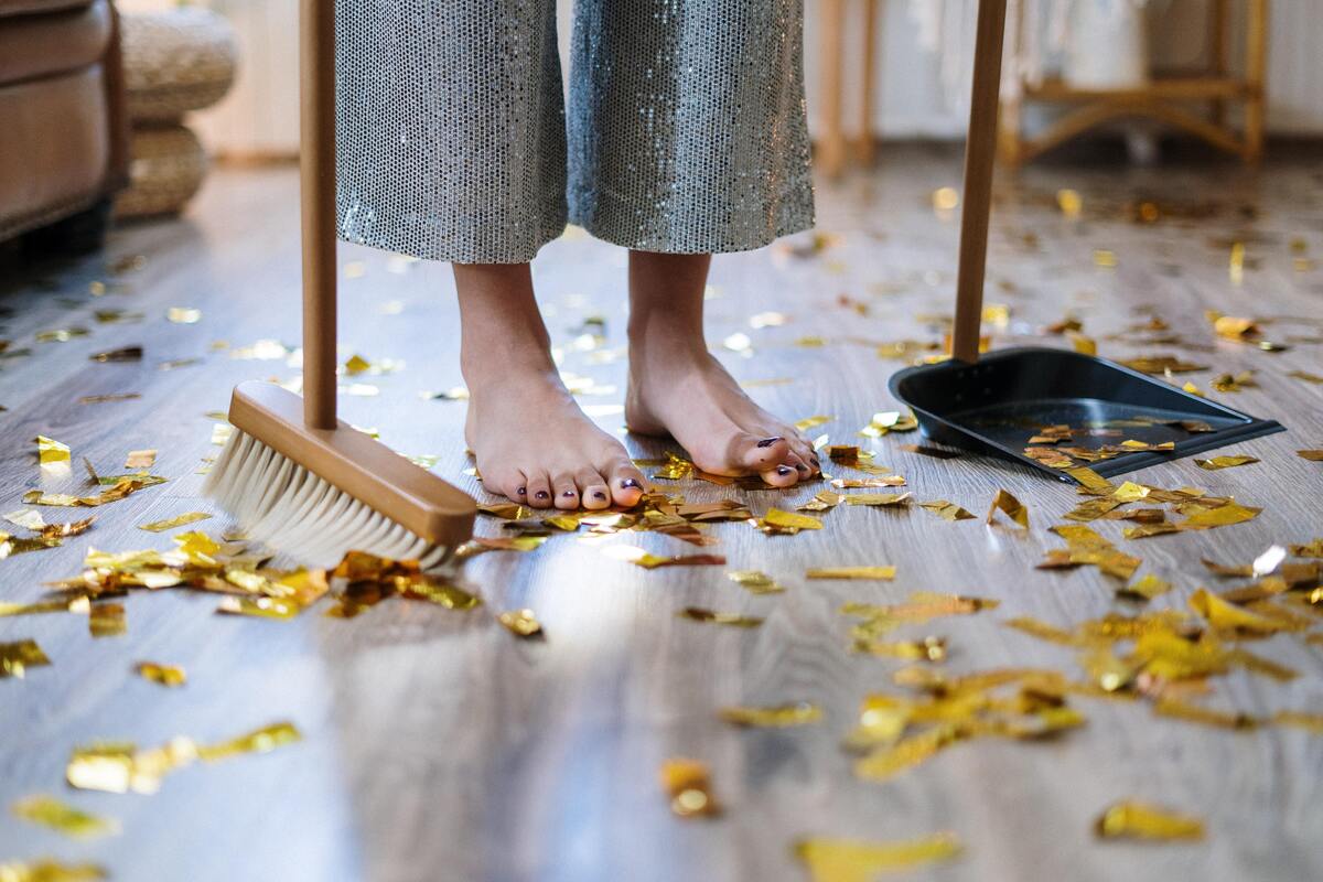 Mulher em pé no chão, segurando vassoura e pá, enquanto o chão está tomado de papéis de festa.