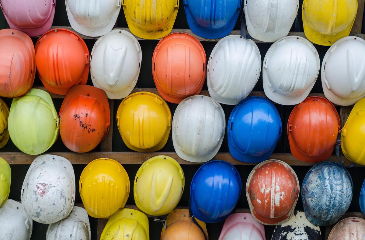 Capacetes de proteção para obras de diversas cores pendurados na parede. 