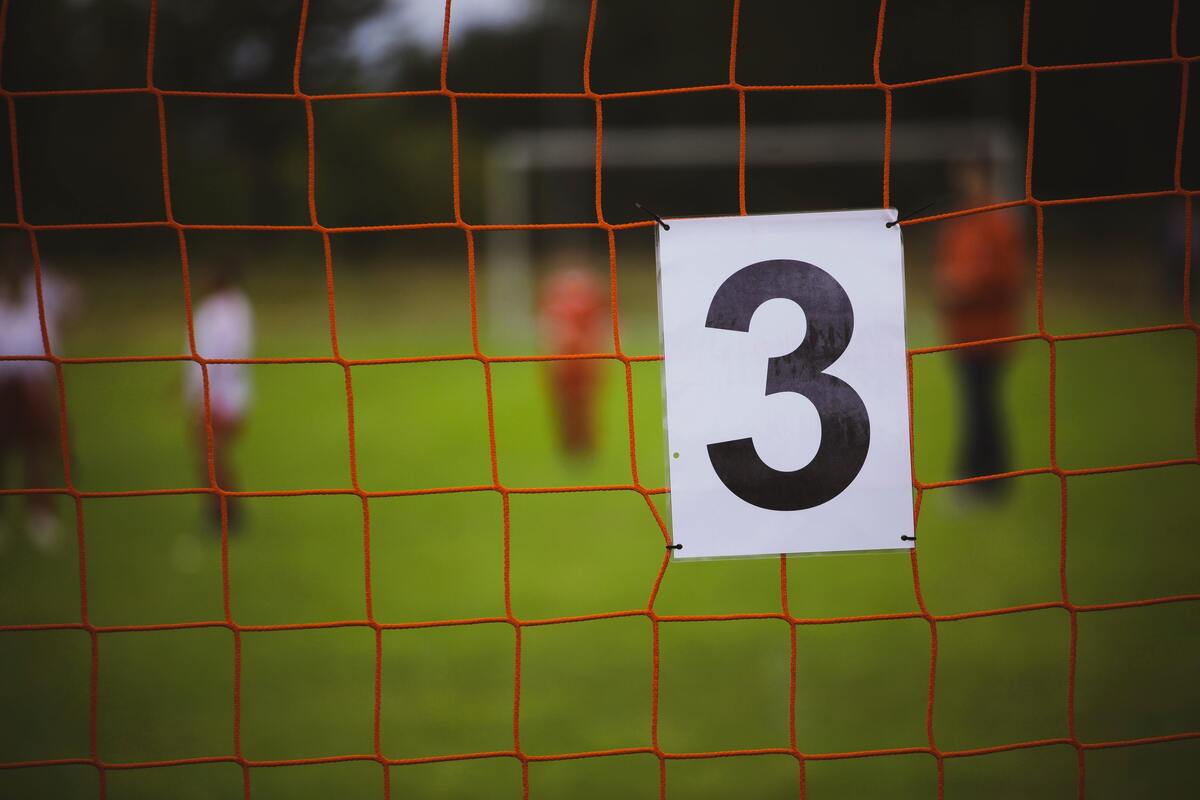 Número três em preso à rede de futebol, time ao fundo desfocado. 