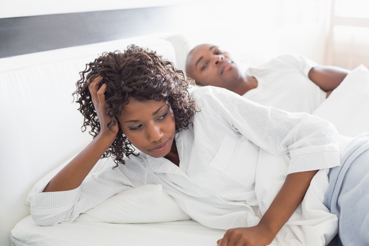 Casal deitado em uma cama. Mulher, com semblante incomodado, olha para o lado oposto ao que o marido está, depois de rejeitá-lo. 