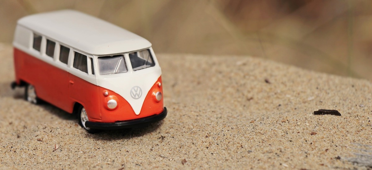 Kombi de brinquedo laranja com detalhes em branco, em cima de monte de areia.