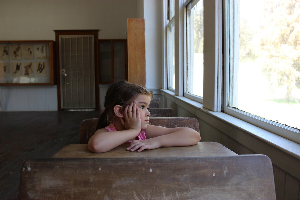 Menina sentada em uma mesa de escola, olhando para a janela.