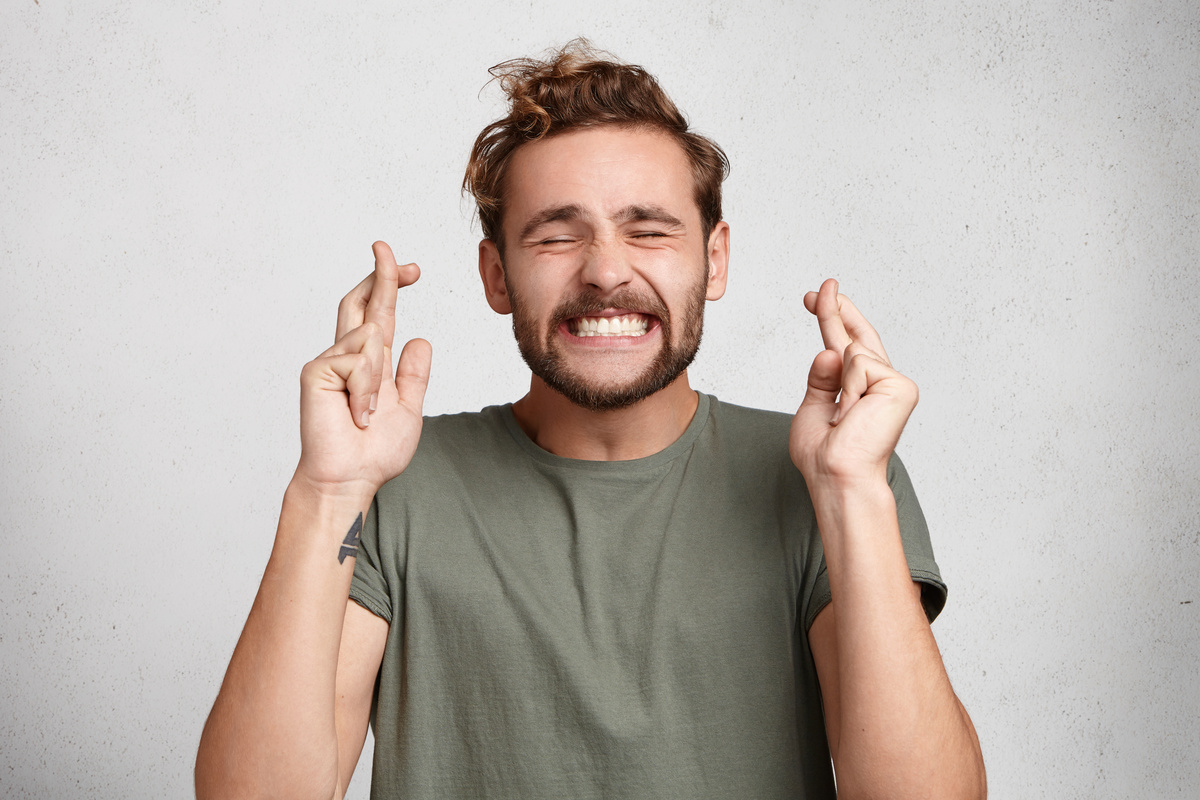 Homem fazendo sinal de figas com os dedos enquanto tem os olhos fechados, representando a procura pela sorte.