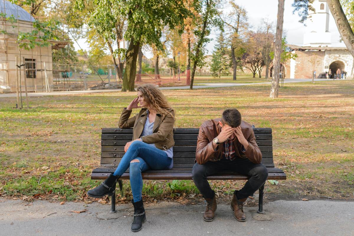 Casal sentado em um banco de praça, olhando em direções opostas com as mãos no rosto.