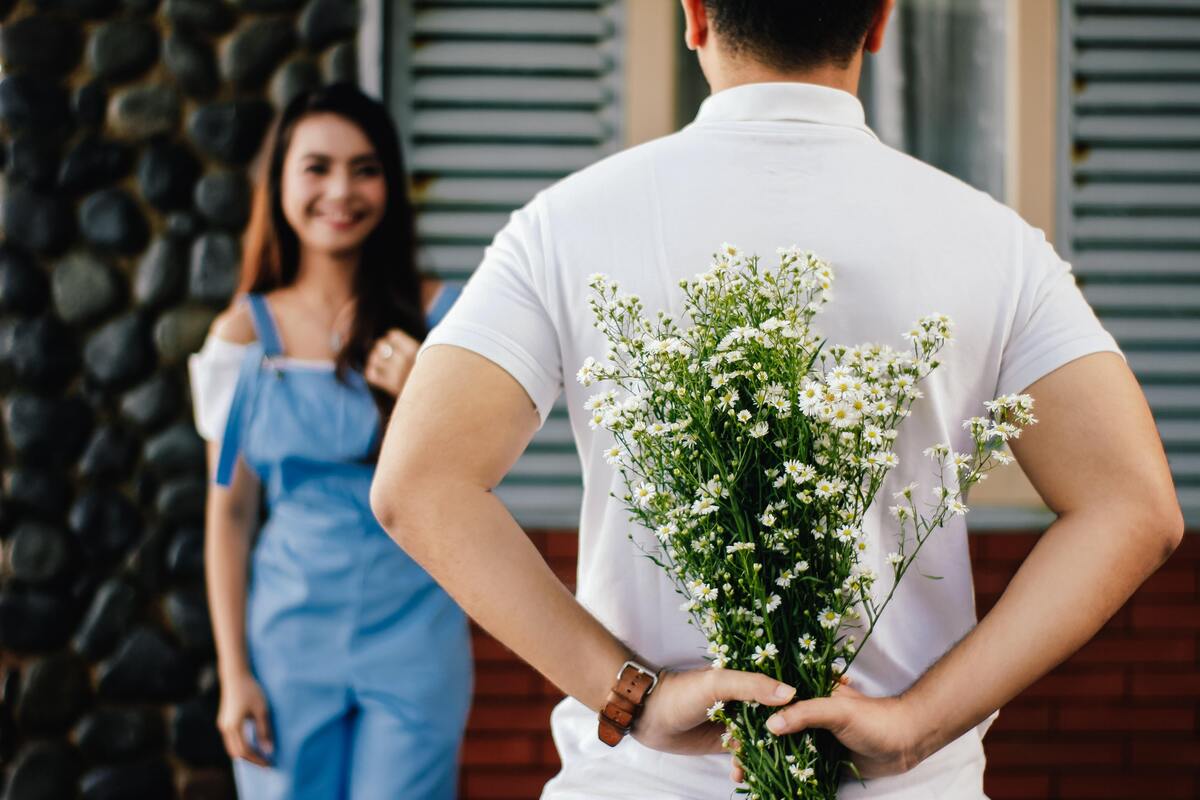 Menina sorri em direção à um homem escondendo flores atrás das costas.