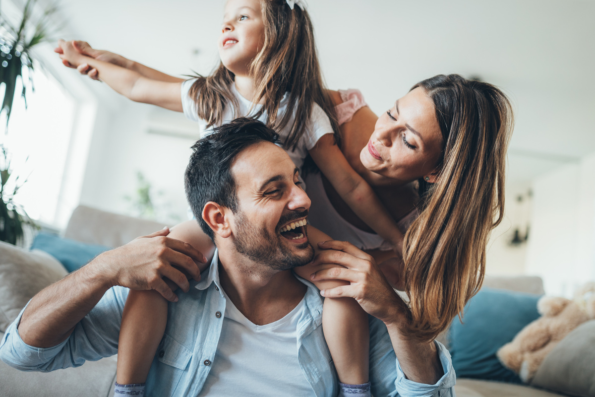 Homem com menina criança montada em suas costas, brincando com ela enquanto sorri para mulher, representando o lado familiar do homem de Câncer.