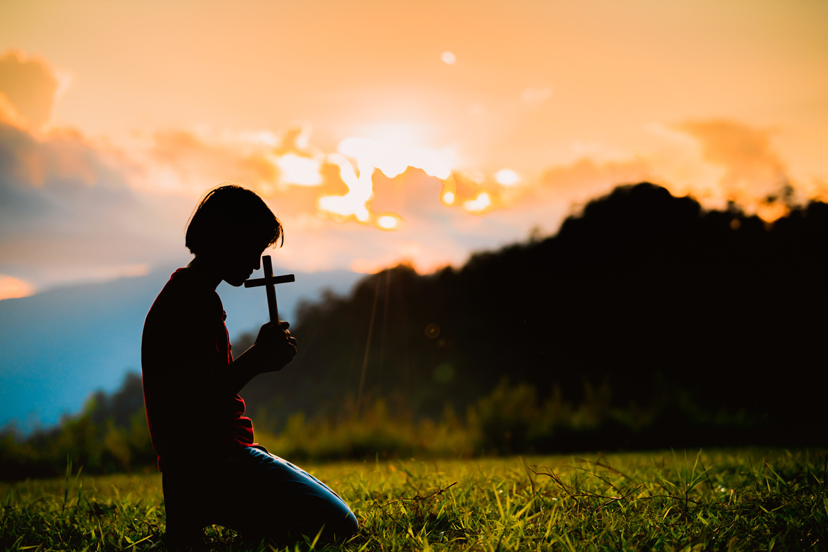 Homem ajoelhado em campo ao por do sol enquanto segura uma cruz pequena em suas mãos, representando a fé trazida pelos salmos.