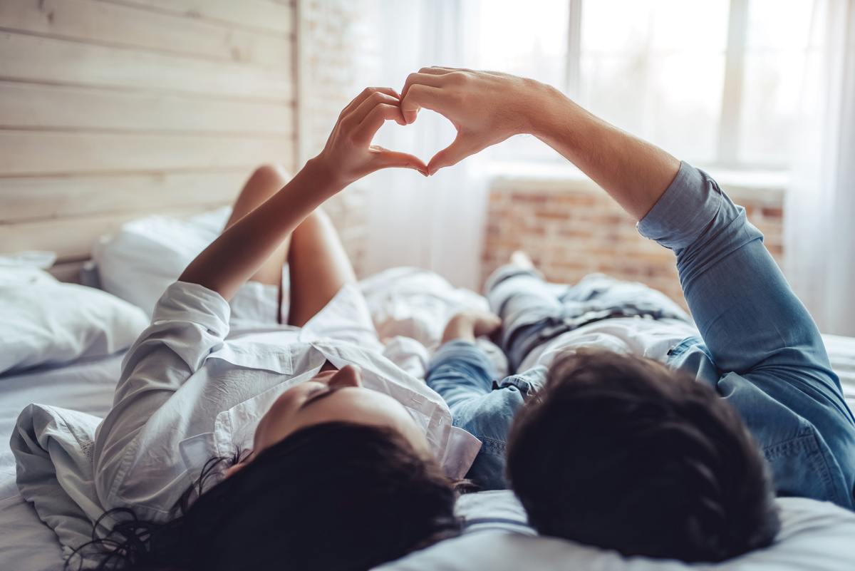 Casal de homem e mulher deitados em cama formando um coração com suas mãos.