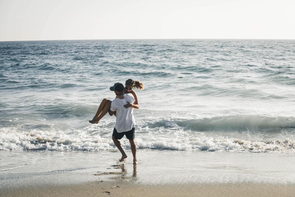 Homem segurando mulher no colo na praia.