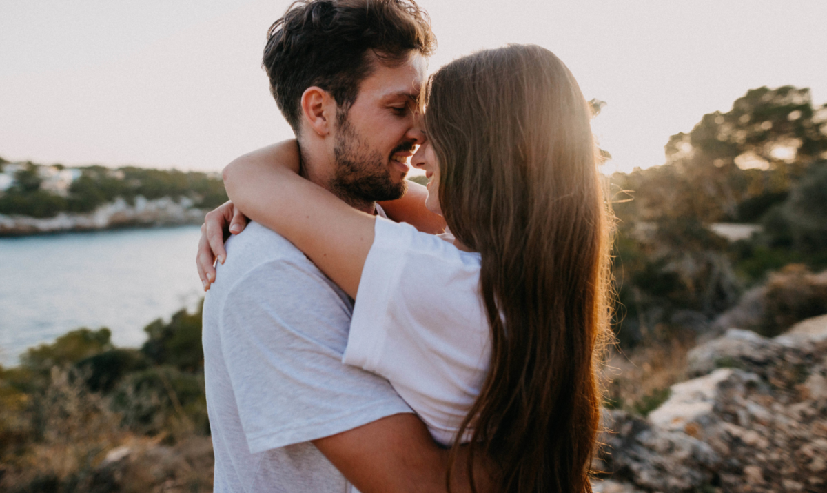 Casal se abraçando, com os narizes se tocando, em cenário com pedras, vegetação e o mar, representando o signo de Peixes no amor.