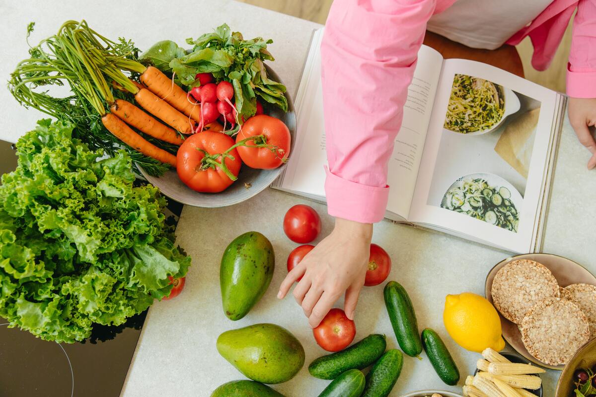 Pessoa com livro de receitas, alface, tomates, pepinos e outros alimentos