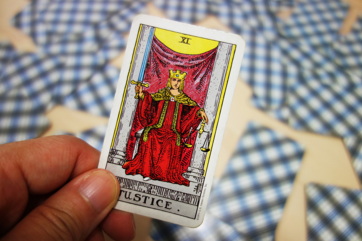 Mão segurando carta A Justiça em frente à mesa com outras cartas de Tarot espalhadas.