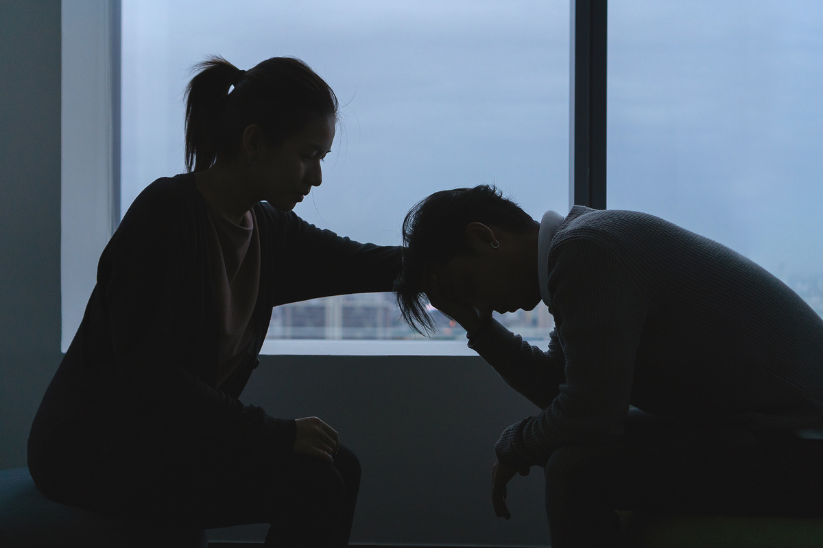 Silhuetas de casal sentado um de frente para o outro, enquanto o homem tem a cabeça baixa e a mulher o conforta, representando o desconforto da amarração amorosa.